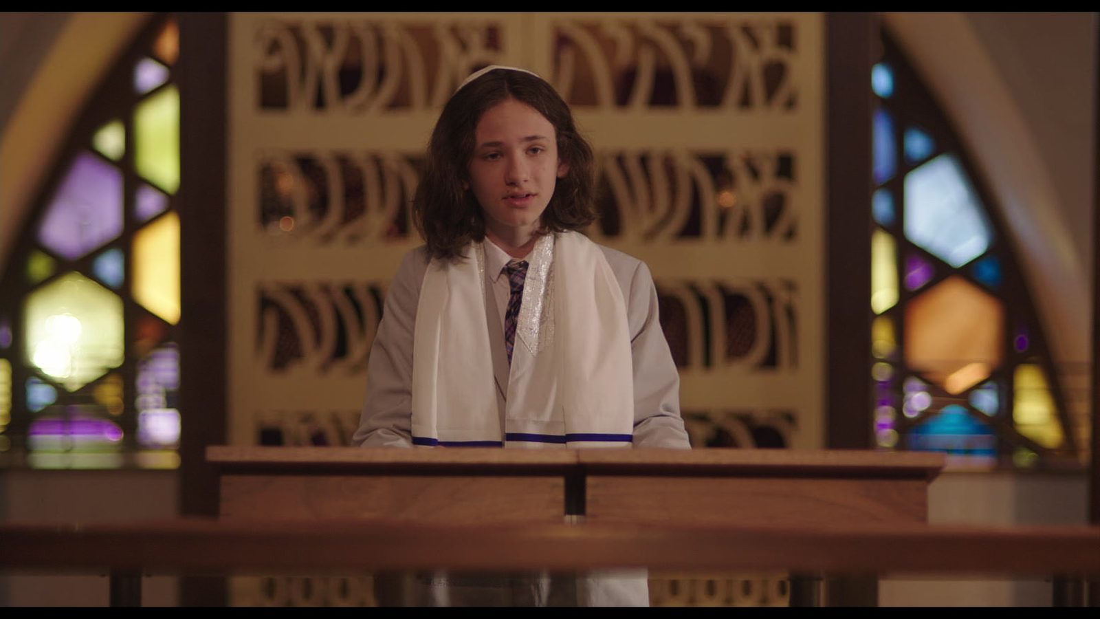 Ethan, in a tallit and kippah, stands at the bimah in front of stained-glass windows